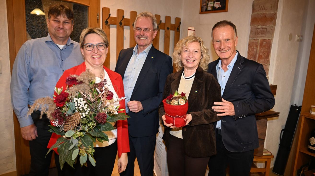 Vier Menschen stehen lächelnd in einem Raum zusammen. Zwei Frauen halten Blumen, eine in Rot und die andere in einer roten Tasche. Zwei Männer stehen hinter ihnen, einer mit einer Plakette. Eine Holzstruktur und ein Bilderrahmen sind im Hintergrund.