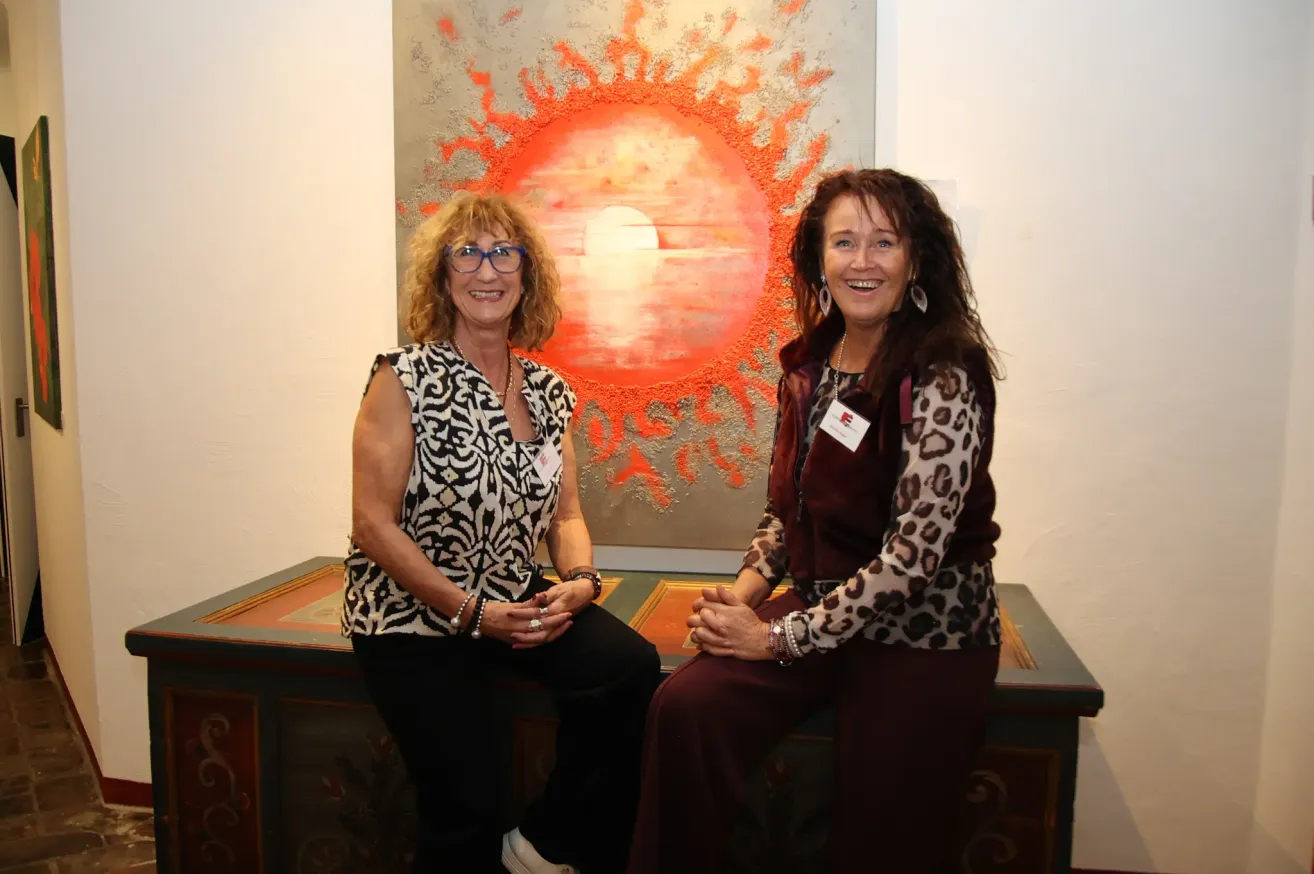 Two women sit next to each other in front of a large painting, smiling and posing for a photo.