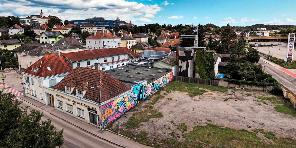 A top view of a European city with various buildings, some with graffiti. A large building has a silo on top. The sky is blue with some clouds.