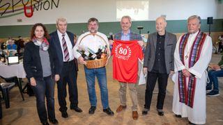 Five men stand together, one holding a red shirt with the number 80 and another holding a basket with bottles. They appear to be celebrating an event.