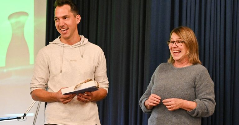 Ein Mann und eine Frau stehen vor einem blauen Vorhang und lächeln. Der Mann hält eine verpackte Schachtel und die Frau hat ihre Hände auf ihrem Bauch.