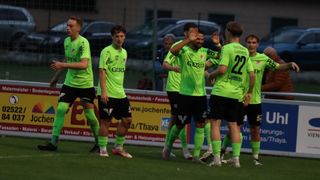 A soccer team wearing green uniforms with numbers and sponsors' logos celebrates on the field, with blurred people and vehicles in the background.