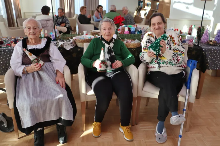 Three elderly women sit in chairs, holding festive items. One woman holds a Christmas tree decoration, while another holds a garland. The room is decorated with Christmas ornaments.
