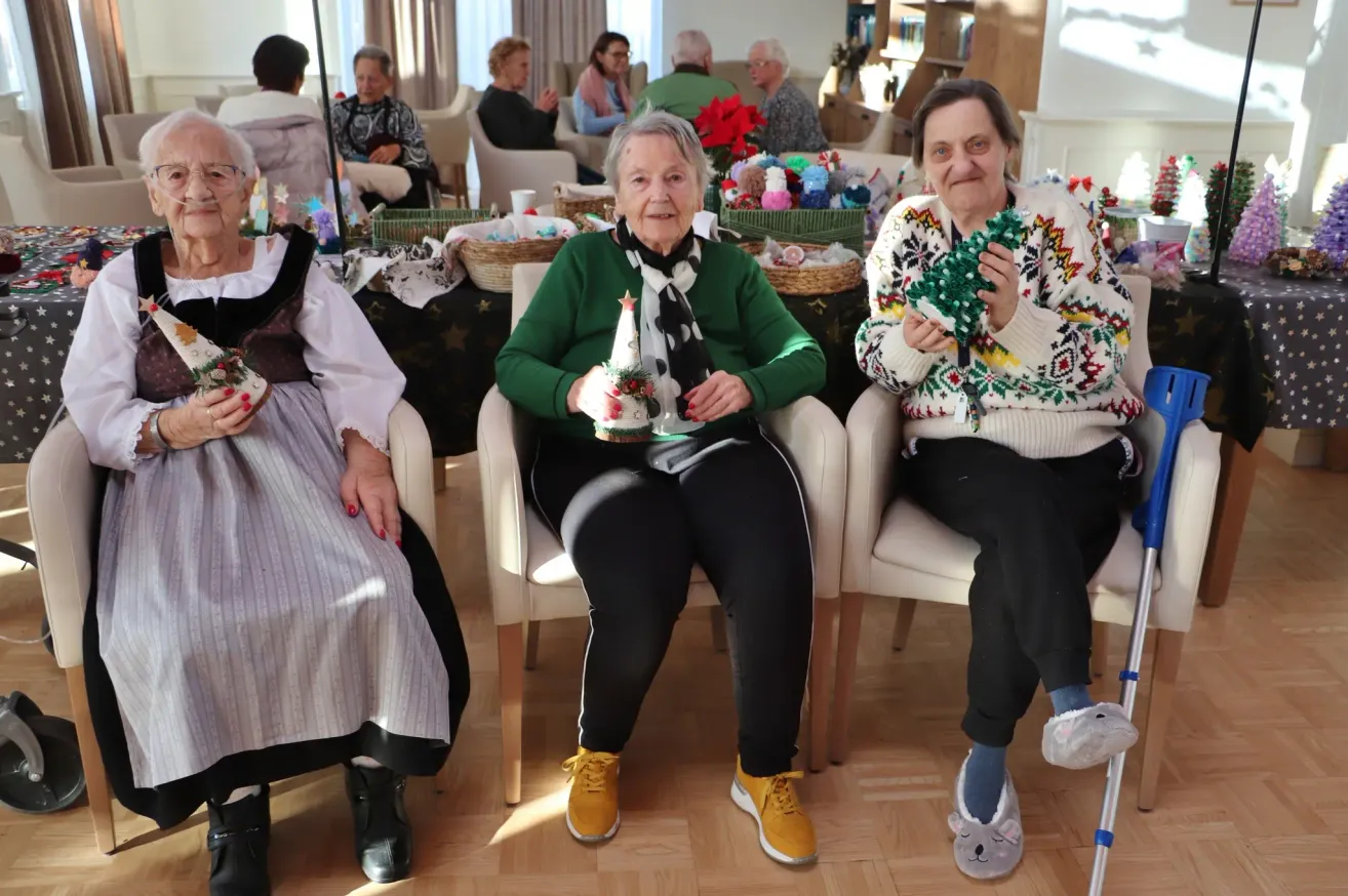 Three elderly women sit in chairs, holding festive items. One woman holds a Christmas tree decoration, while another holds a garland. The room is decorated with Christmas ornaments.