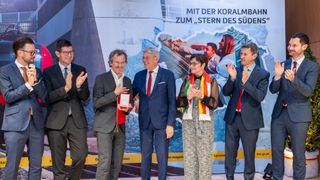 Five individuals in formal attire are standing on a stage, clapping. A banner behind them has a collage of images and text.