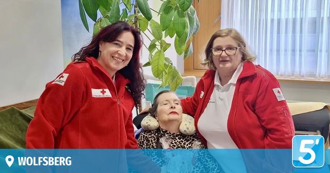 Three women are smiling in a room, two wearing red jackets and one in a leopard print shirt. A large plant and a window with white curtains are in the background. The image has a watermark of 5'Wolfsberg'.