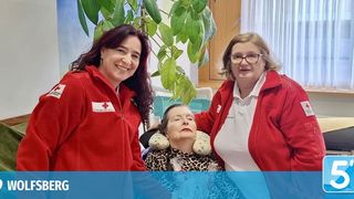 Three women are smiling in a room, two wearing red jackets and one in a leopard print shirt. A large plant and a window with white curtains are in the background. The image has a watermark of 5'Wolfsberg'.