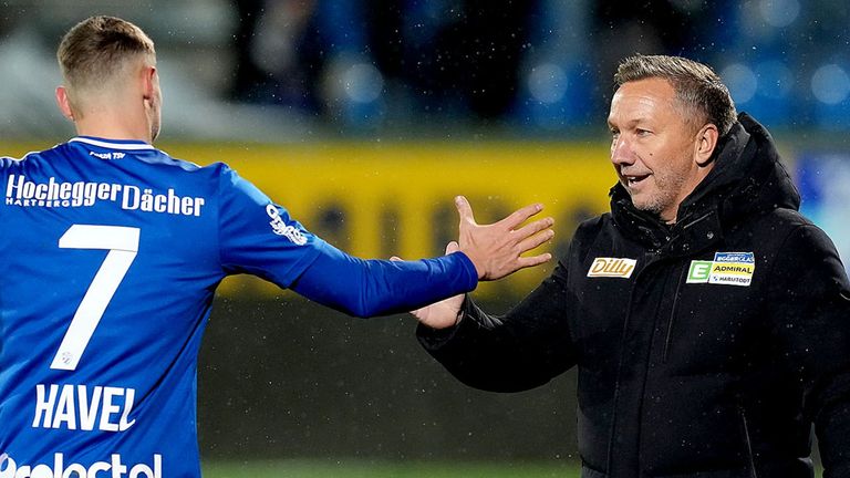 A man in a blue shirt shakes hands with another man in a black jacket, likely in a sports event. The man in blue wears a logo on his sleeve.