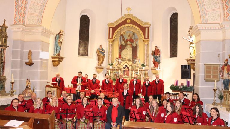 Eine große Gruppe von Musikern in roten Uniformen posiert für ein Foto in einer Kirche. Ein Mann in einem schwarzen Mantel sitzt vorne. Dahinter ist ein großer Altar mit Statuen geschmückt.