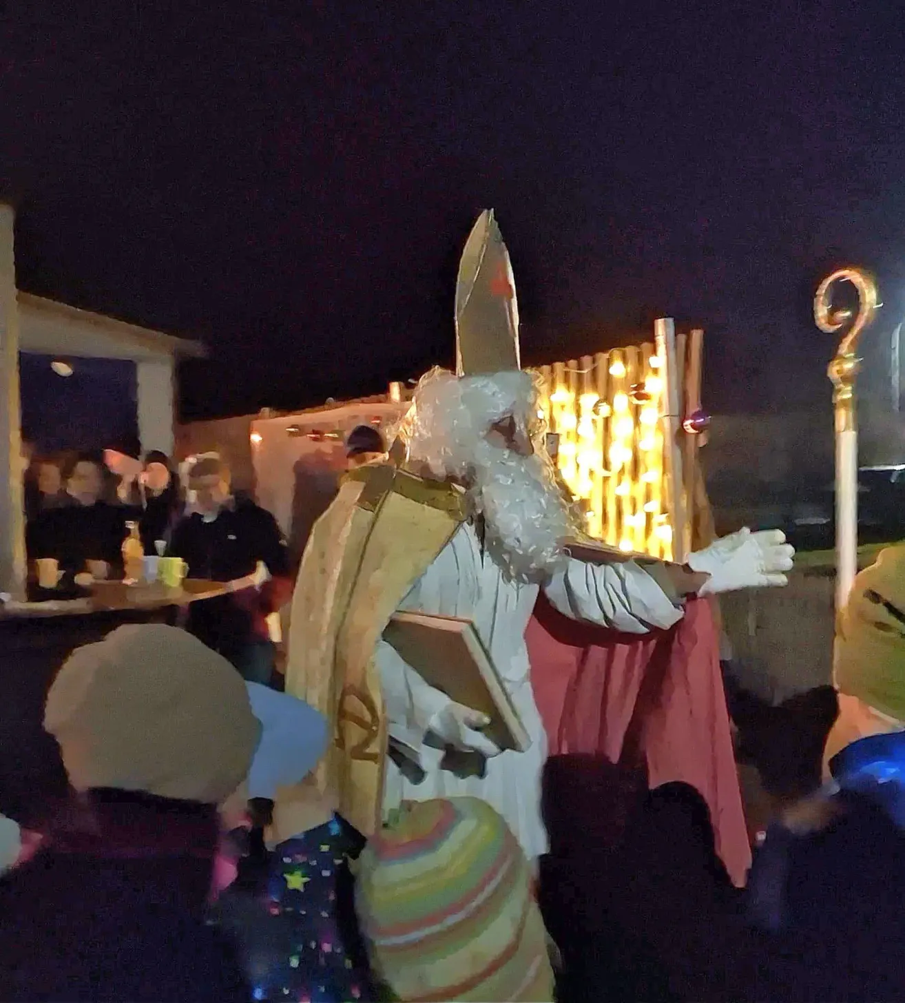 Eine Person, die als St. Nikolaus verkleidet ist, unterhält eine Menschenmenge bei Nacht. Die Menge besteht hauptsächlich aus Kindern, die Mützen tragen, und einigen Erwachsenen. Im Hintergrund befindet sich ein Tisch mit Tassen und ein Gebäude mit Lichtern.