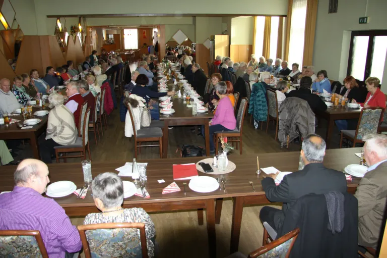 Eine große Gruppe älterer Menschen speist in einem Saal. Die Tische sind mit Tellern, Besteck und Gläsern gedeckt. Einige Menschen tragen Brillen und Jacken. Der Saal hat Holzböden und Wände mit Vorhängen und Bilderrahmen.