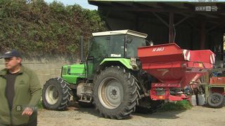A green tractor with a red attachment is parked in front of a barn. The tractor has a license plate with the number 4971.