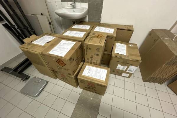 A bathroom with a sink, boxes, and a weighing scale. The boxes are stacked on the tiled floor. Each box has a label on it.