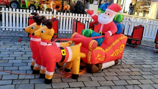An inflatable Santa Claus sits in his sleigh, pulled by two inflatable reindeer on a brick sidewalk. Behind them is a white fence and a horse-drawn carriage.