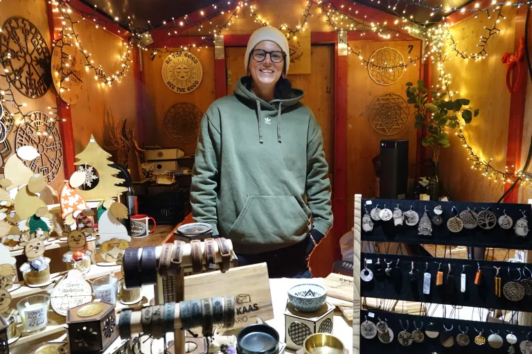 A smiling woman in a green hoodie stands behind a display table with various items, including bracelets, cups, and bowls, in a room decorated with Christmas lights and ornaments.