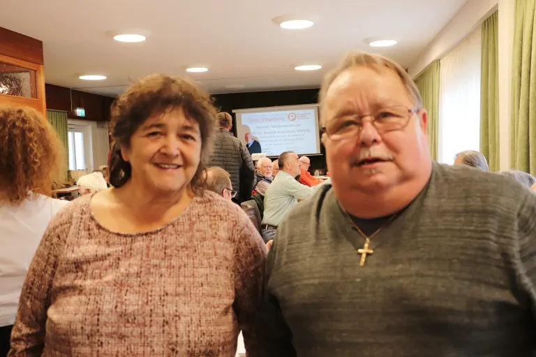 Eine lächelnde Frau und ein Mann mit Brille stehen gemeinsam im Innenbereich, mit einer Projektionsleinwand, die eine Willkommensnachricht hinter ihnen anzeigt. Andere Menschen sitzen im Hintergrund an Tischen.