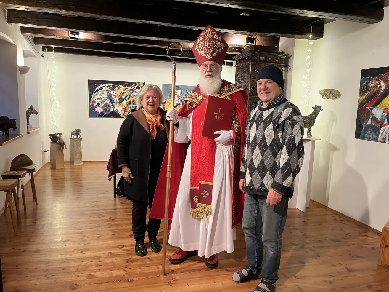 Ein Mann, der als Bischof verkleidet ist, steht zwischen zwei lächelnden Erwachsenen in einem Raum mit Holzböden und weißen Wänden.