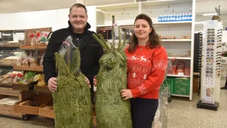 A man and a woman stand in a store, each holding a large artificial Christmas tree. They are smiling, with shelves and products in the background.