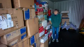 An elderly woman stands in a room filled with numerous gift boxes, wrapped in colorful paper. She is wearing a green jacket and jeans, and appears to be looking at the camera.