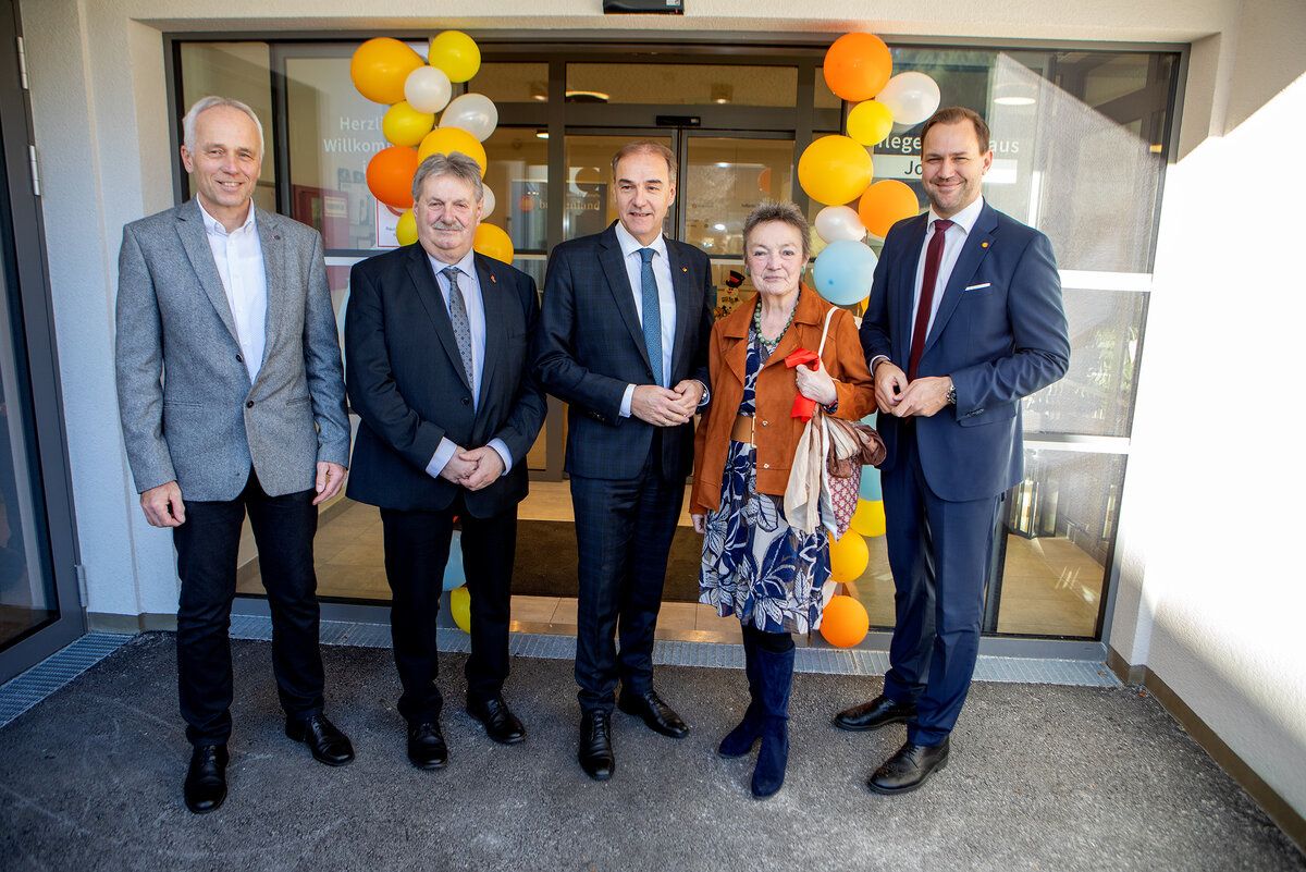 Five individuals stand in front of a building entrance, wearing formal attire. The woman in the center holds a red object. Balloons are visible behind them.