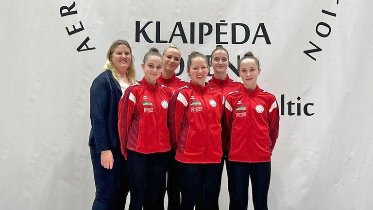 Fünf Frauen in roten Jacken posieren für ein Foto vor einem weißen Banner mit dem Wort Klaipeda. Sie lächeln und schauen in die Kamera.