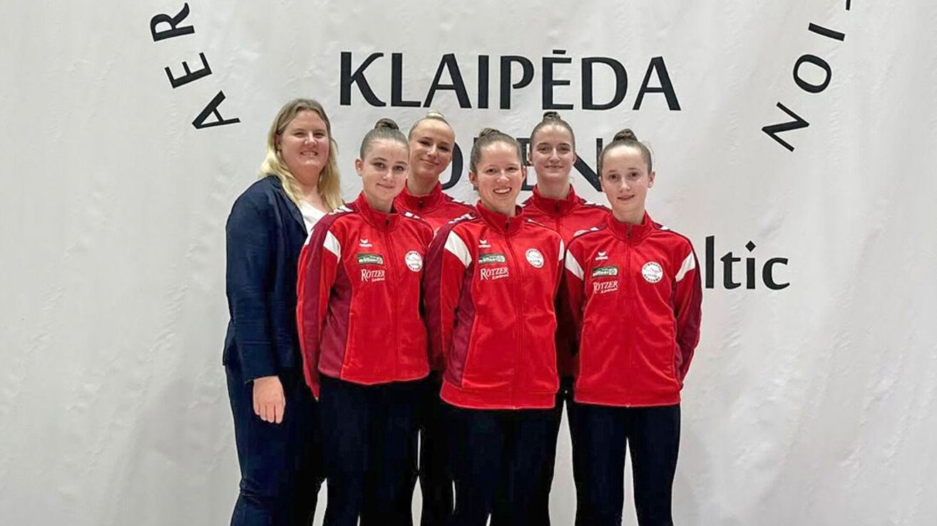 Fünf Frauen in roten Jacken posieren für ein Foto vor einem weißen Banner mit dem Wort Klaipeda. Sie lächeln und schauen in die Kamera.