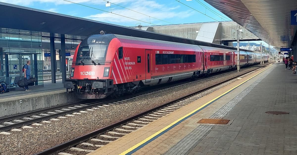 Ein roter Zug steht auf einem Bahnsteig, der Text 'Heute. Für morgen. Für uns.' ist zu sehen. Der Zug hat eine glänzende Außenseite und steht unter einem Dach. Der Bahnsteig hat einen Ziegelboden mit einer gelben Linie.
