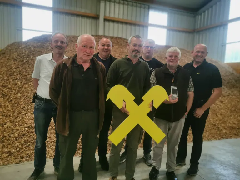 Sieben Männer stehen in einem Lagerhaus und lächeln für ein Foto. Sie tragen Freizeitkleidung, und einer hält einen Preis. Sie sind von einem Haufen Holzspäne umgeben.