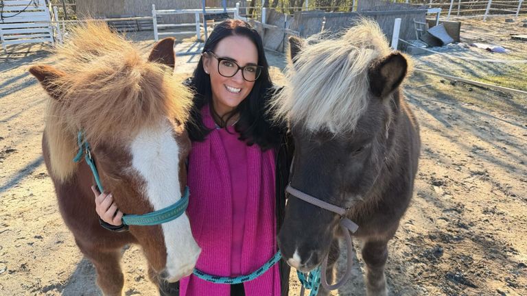 Eine lächelnde Frau mit Brille posiert mit zwei Pferden, eines mit einem braun-weißen Fell und das andere mit einem schwarz-weißen Fell.