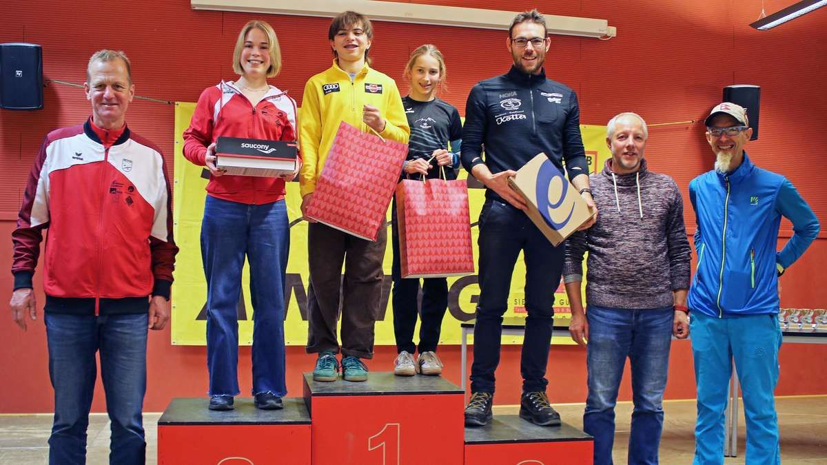 Fünf Personen stehen auf einem Podest mit nummerierten Stufen, wahrscheinlich bei einer Preisverleihung. Drei Personen sind auf der obersten Stufe, jede hält einen Geschenkbeutel. Die linke Person hält eine Saucony-Box. Dahinter zeigt ein Banner Buchstaben und Zahlen.