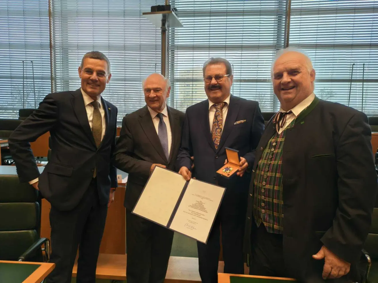 Vier Männer in formellen Anzügen stehen in einem Raum und lächeln für ein Foto. Ein Mann hält ein gerahmtes Dokument, und ein anderer hält eine Medaille. Der Raum hat Holzbänke und Fenster mit Jalousien.