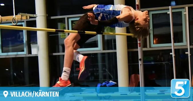 Ein junger Athlet führt einen Hochsprung in einer Indoor-Sportanlage durch. Er trägt ein blaues und weißes Trikot und Turnschuhe, und sein Kopf ist nach hinten geneigt.