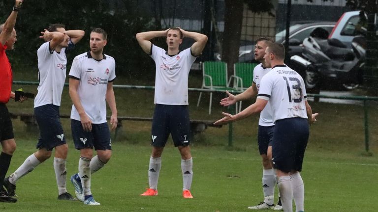Fußballspieler in weißen Trikots stehen auf einem Rasenfeld im Regen. Ein Spieler trägt die Nummer 7 und hat die Hände im Nacken. Dahinter schauen andere zu. Grüne Stühle und Autos sind im Hintergrund.