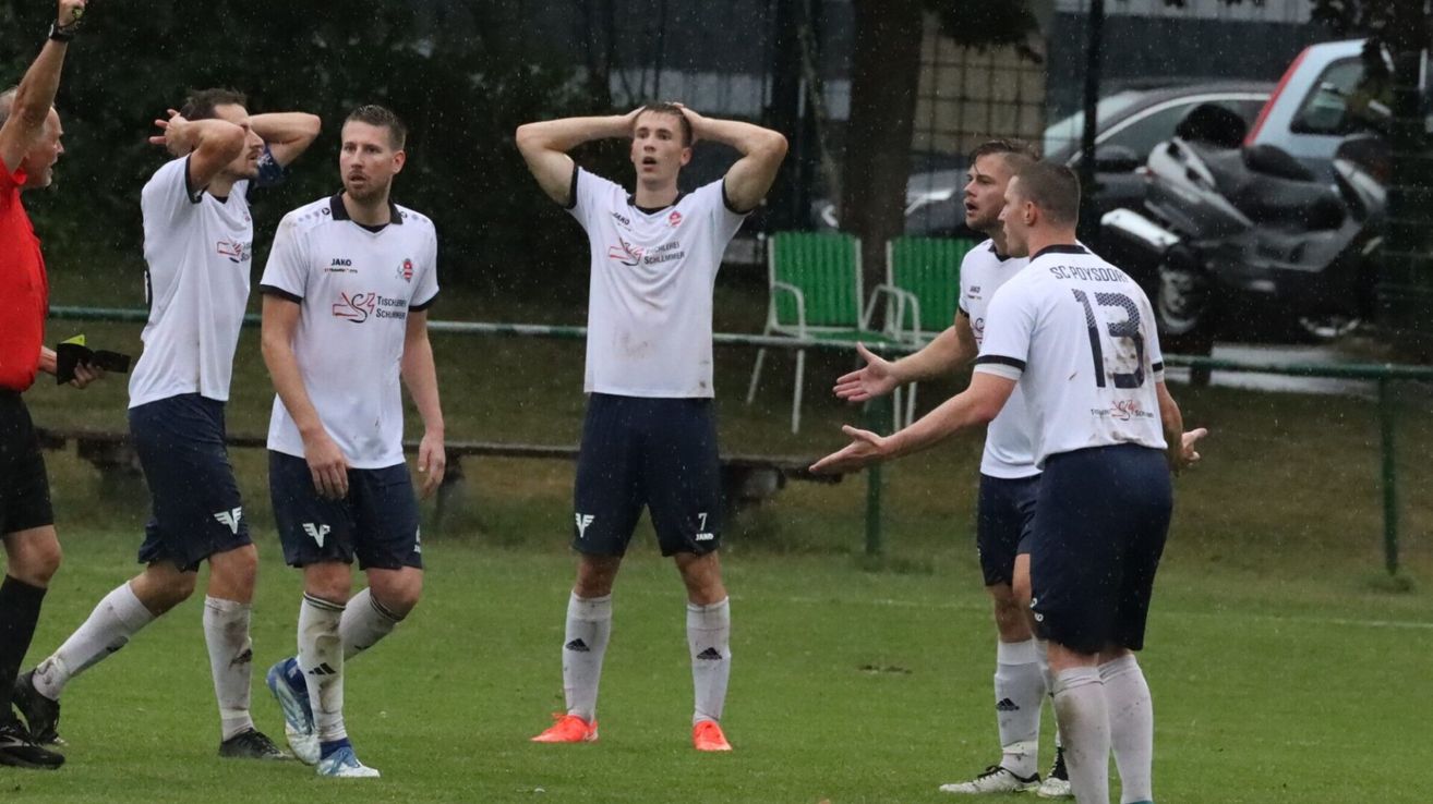 Fußballspieler in weißen Trikots stehen auf einem Rasenfeld im Regen. Ein Spieler trägt die Nummer 7 und hat die Hände im Nacken. Dahinter schauen andere zu. Grüne Stühle und Autos sind im Hintergrund.