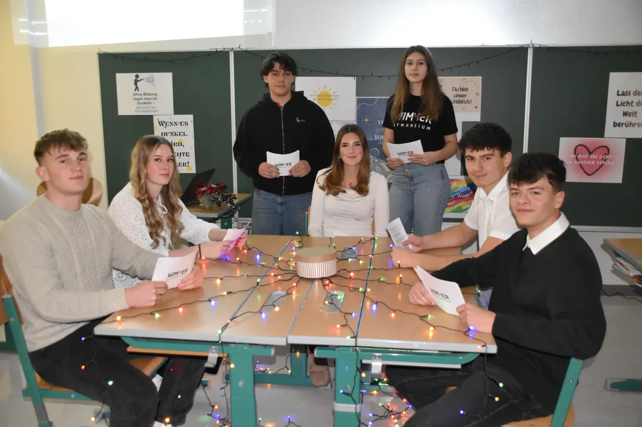 Eine Gruppe von Menschen sitzt um einen Tisch mit Weihnachtslichtern. Sie halten Papiere und lächeln, möglicherweise für ein Foto. Eine Frau steht hinter ihnen, und Plakate mit Botschaften hängen an der Wand.