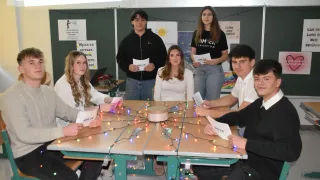 A group of people sit around a table with Christmas lights. They hold papers and smile, possibly for a photo. A woman stands behind them, and posters with messages are on the wall.