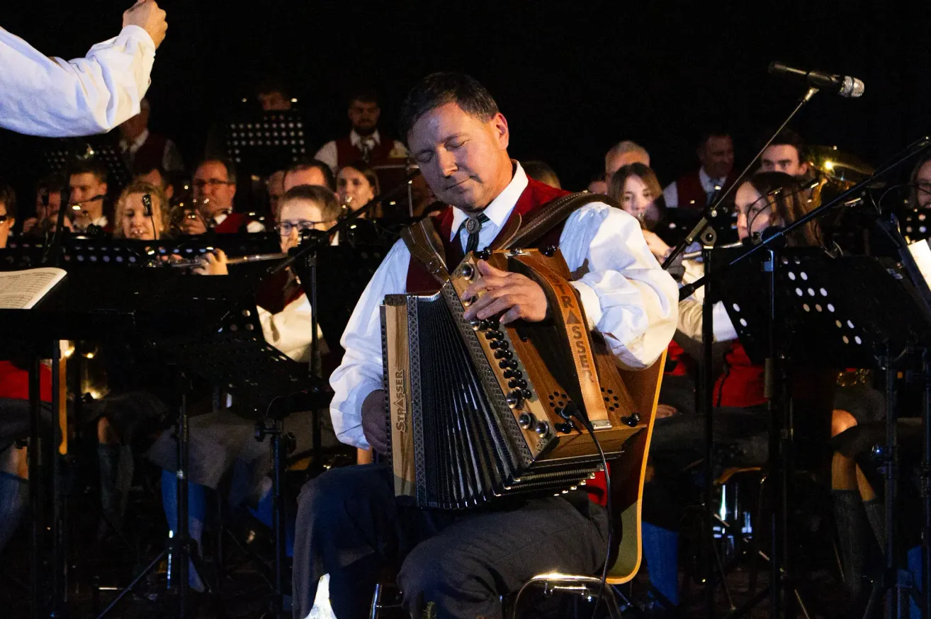 A man plays an accordion in a concert with an orchestra behind him, some wearing glasses and holding instruments.