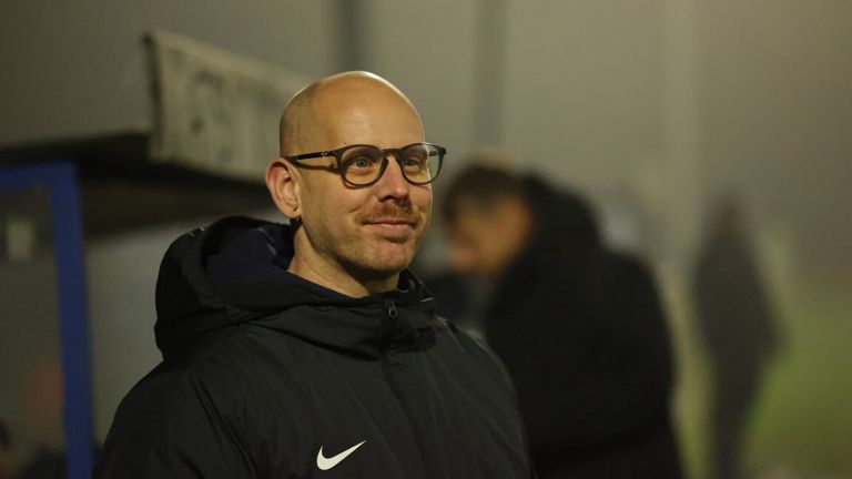 Ein kahlköpfiger Mann mit Brille, der eine schwarze Nike-Jacke mit einem weißen Nike-Logo auf der linken Seite trägt, steht vor einem verschwommenen Hintergrund.