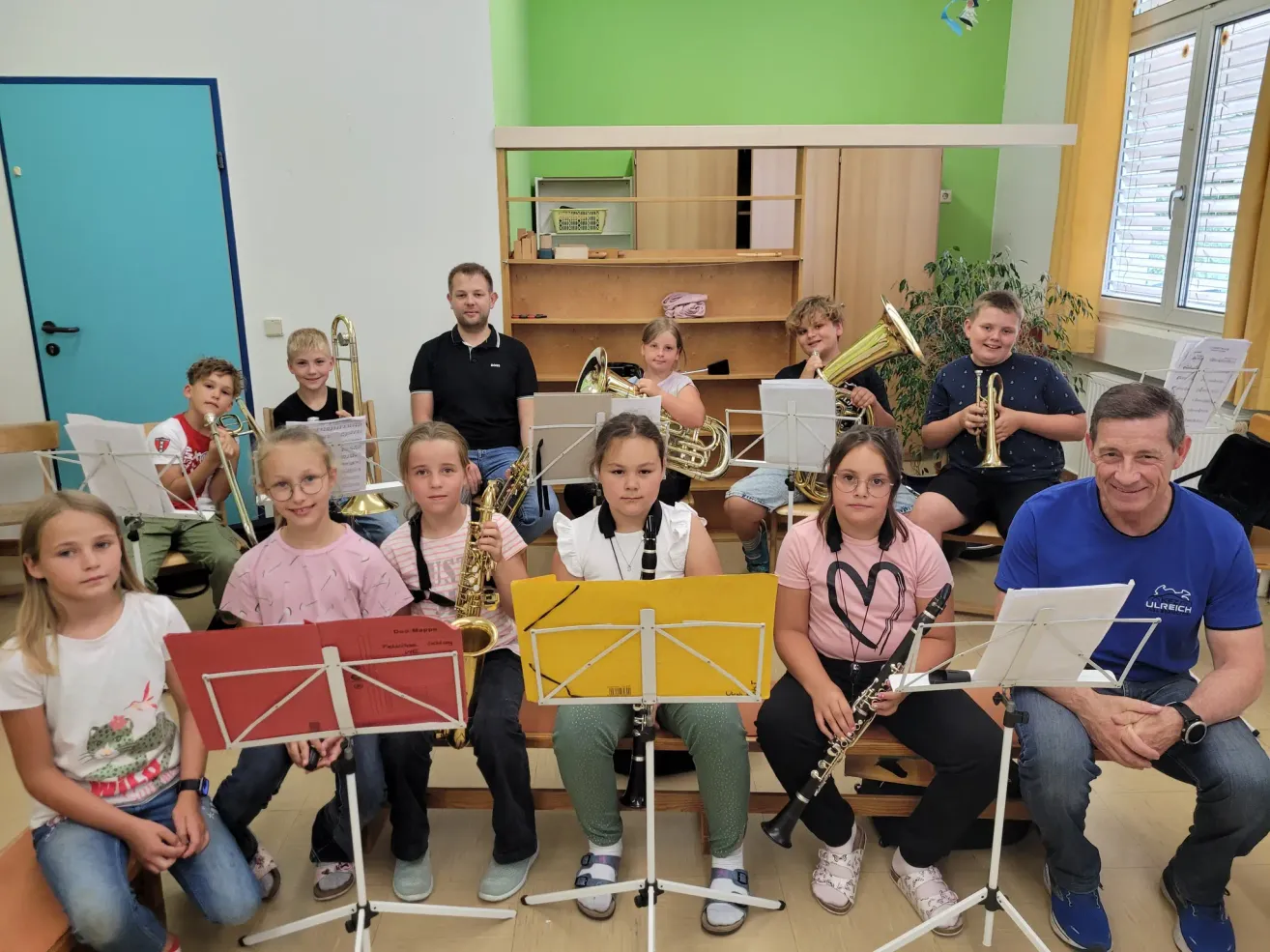 Ein Klassenzimmer mit Schülern und Lehrern. Eine Gruppe von Kindern spielt Musikinstrumente, darunter Trompeten, Saxophone und eine Klarinette. Einige Erwachsene sind anwesend und sitzen auf der Bank. Der Raum hat eine grüne Wand und ein Holzregal.