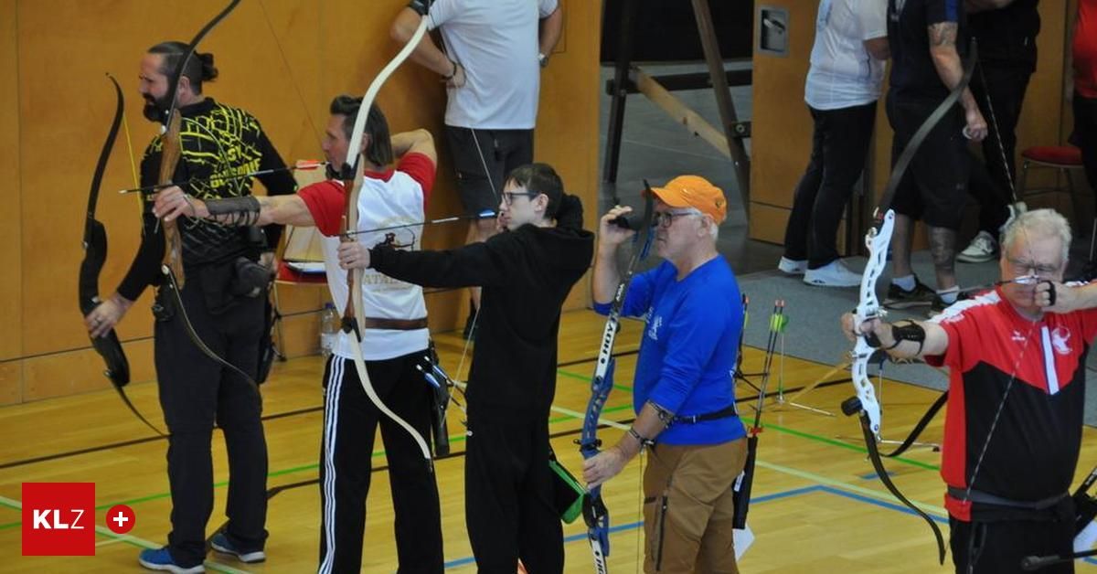Eine Gruppe von Menschen übt Bogenschießen in einer Turnhalle. Eine Person hält einen Bogen, während eine andere Person durch ein Fernglas schaut.