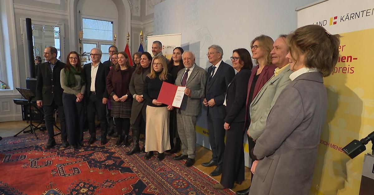Eine Gruppe von Menschen in formeller Kleidung steht drinnen und hält eine rote Mappe. Eine Frau in der Mitte hält die Mappe und ein Mann neben ihr lächelt. Hinter ihnen ist eine weiße Wand mit Text.