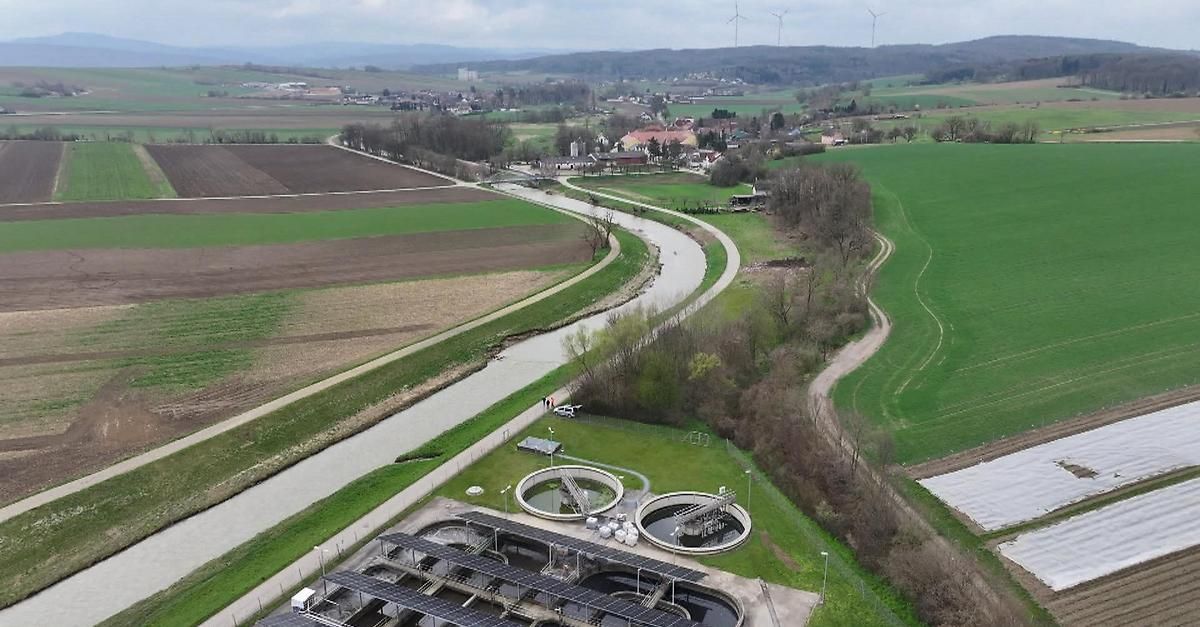 Luftaufnahme einer Kläranlage mit Solarmodulen. Die Anlage ist von grünen Feldern und einem mäandernden Fluss umgeben. Gebäude und Windräder sind in der Ferne sichtbar.