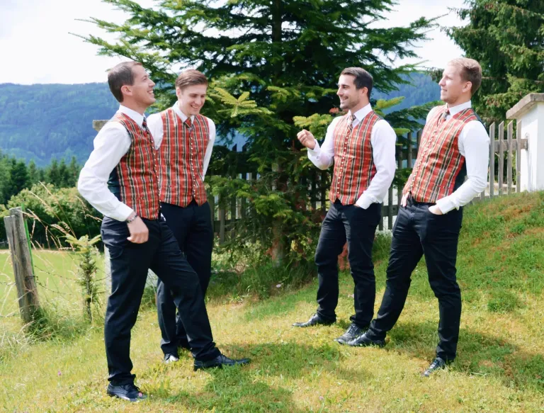 Vier Männer in roten und schwarzen Westen und passenden Hosen stehen in einem Grasbereich, lächeln und schauen sich gegenseitig an. Sie scheinen für ein Foto zu posieren.