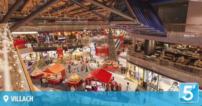 Ein Blick von oben auf ein geschäftiges Einkaufszentrum, geschmückt mit Weihnachtsdekorationen, mit einem zentralen Weihnachtsbaum, Ständen und Menschen, die herumlaufen. Das Einkaufszentrum hat mehrere Ebenen mit Geschäften und Sitzbereichen.