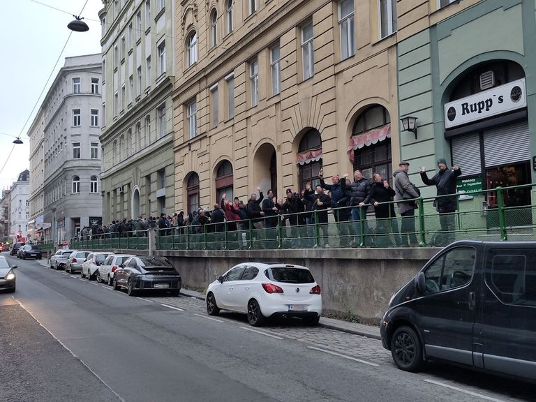 Eine Menschenmenge steht auf einem Balkon über einer Straße und beobachtet die an der Straße stehenden Autos. Der Balkon hat ein grünes Geländer. Ein Van und ein Auto sind in der Nähe geparkt.