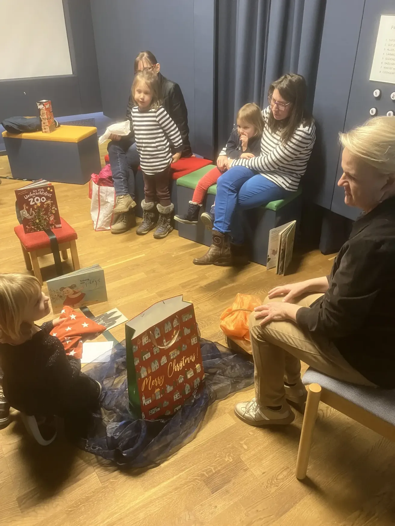In einem Raum sitzen mehrere Kinder um einen Erwachsenen herum, möglicherweise in einem Wartebereich. Einige Kinder haben Bücher, während andere Taschen haben. Ein Kind hat einen roten Weihnachtsbeutel.