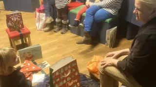 In a room, several children are seated around an adult, possibly in a waiting area. Some children have books, while others have bags. One child has a red Christmas bag.