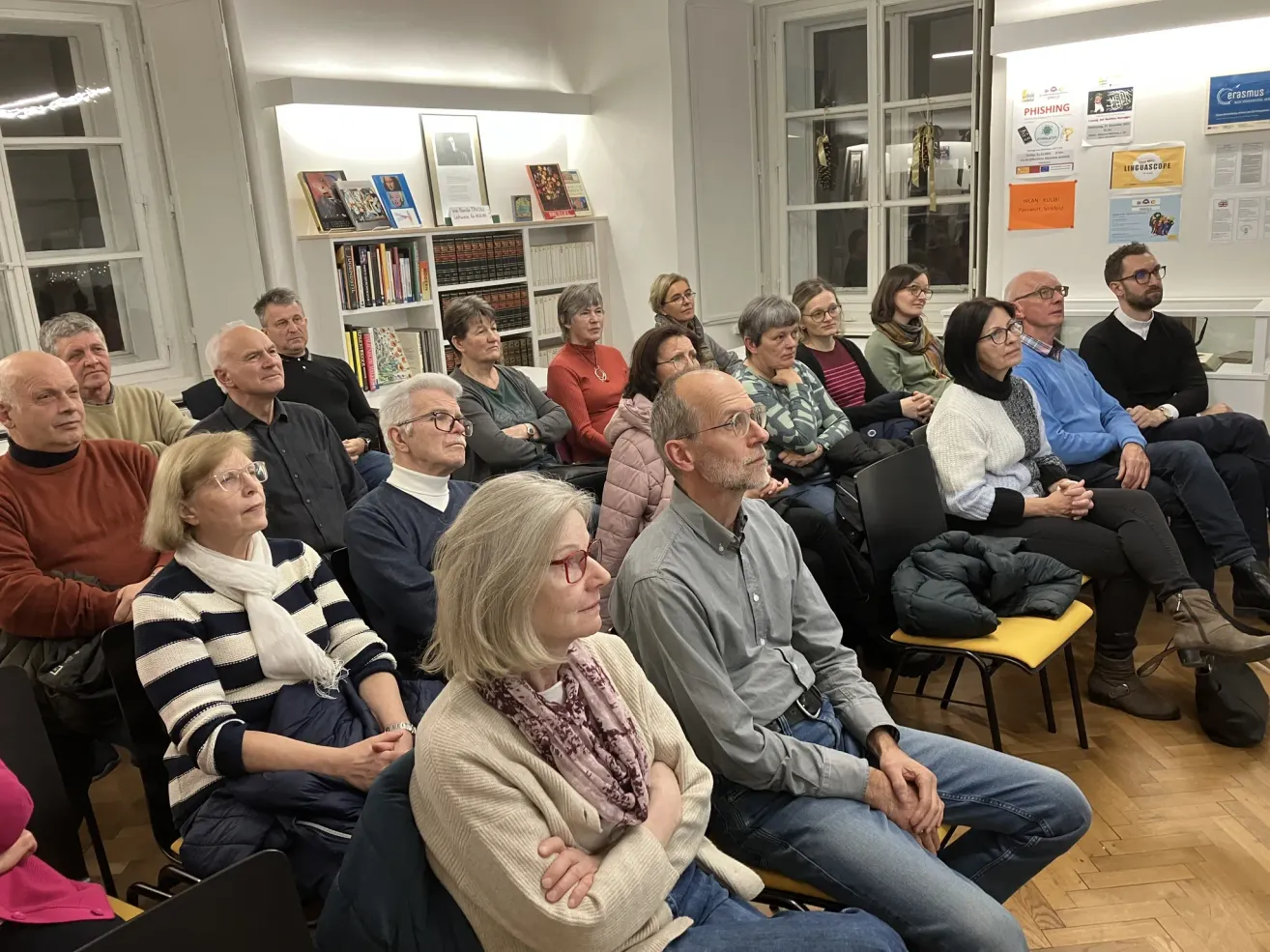 Eine Gruppe von Menschen sitzt in einem Raum mit Bücherregalen und einem Schild. Einige tragen Brillen und andere Schals.