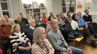 A group of people are sitting in a room with bookshelves and a sign. Some are wearing glasses and some are wearing scarves.