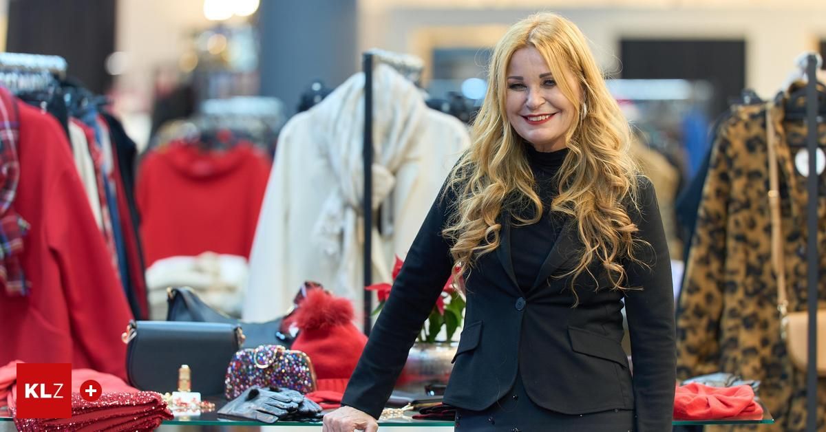 Eine Frau mit blonden Haaren steht lächelnd vor einem Bekleidungsgeschäft, in einem schwarzen Anzug. Hinter ihr hängen Kleidungsstücke, und auf einem Glastisch sind Accessoires.